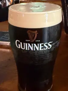 A pint glass of Guinness stout beer with a creamy head, sitting on a wooden surface. The Guinness logo with a harp is visible on the glass.