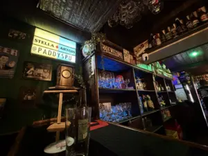 A dimly lit bar with shelves stocked with bottles and glassware, neon signs, and eclectic decor. A glass clock and a Stella Artois sign with "@PADDYSPU" are visible on the wall above the bar counter.