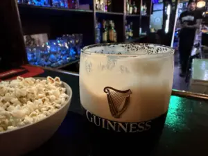 A pint of Guinness stout sits on a bar next to a bowl of popcorn, with shelves of glasses and bottles in the background and a person walking in the distance.