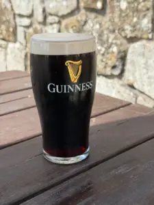 A pint glass of Guinness stout with a creamy head sits on a wooden outdoor table, with a stone wall in the background on a sunny day.