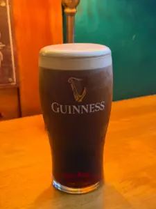 A Guinness pint glass with a creamy white head sits on a wooden table in a warmly lit pub setting.