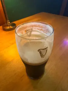 A half-full Guinness pint glass with a creamy head sits on a wooden table next to a brass lamp in a cozy pub. The glass features the distinctive harp logo.