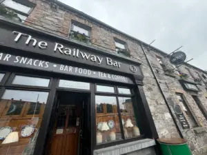 The exterior of The Railway Bar, a stone building with flower boxes and large windows, features signs for snacks, bar food, tea, coffee, and a guinness pint glass pub on a cloudy day.