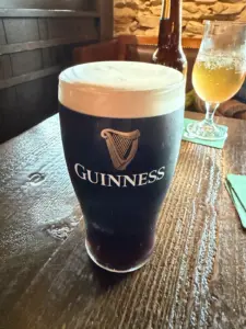 A Guinness pint glass with a creamy head sits on a wooden table in a pub, with another glass and a bottle in the background.
