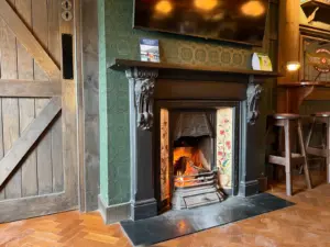 A cozy pub interior with a lit fireplace featuring ornate black detailing, patterned tiles, green wallpaper, and wooden furniture—including a round table set with a Guinness pint glass on the parquet floor.