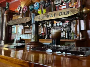A close-up of a bar counter with brass "Railway Bar" tap handles, various taps, and a pint in a Guinness glass. Shelves with bottles and glasses are visible in the background of this inviting pub scene.