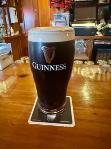 A Guinness pint glass rests on a coaster atop a polished wooden bar counter in a cozy pub, with shelves and glassware visible in the background.