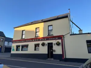 A cream and red two-story pub named "The Railway Bar" with hanging flower baskets, large windows, and a sign from 1860. A Guinness pint glass is visible in the evening sunlight as the building stands under a clear blue sky.