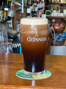 A Guinness pint glass with a creamy head sits on a green coaster atop a wooden bar in a cozy pub, with glasses and snacks visible in the blurred background.