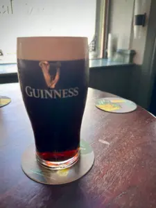 A pint of Guinness with a creamy head sits on a coaster on a wooden pub table, with sunlight streaming through a nearby window. Other coasters are visible on the table.