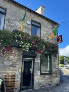 A stone pub building with flower boxes and two green and yellow flags above the entrance. A Guinness sign hangs outside. The sky is blue with some clouds, and there's a barrel by the door.