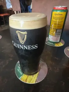 A pint of Guinness stout with a creamy head sits on a coaster on a dark table beside a can of cider. The Guinness logo is visible on the glass.