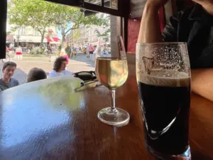 A close-up of a pub table with a glass of white wine and a pint of dark beer. People are sitting outside in a sunny, lively square seen through an open window.