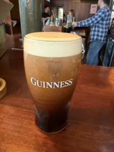 A pint of Guinness beer with a creamy foam head sits on a wooden bar counter, with bartenders and bar equipment visible in the background.