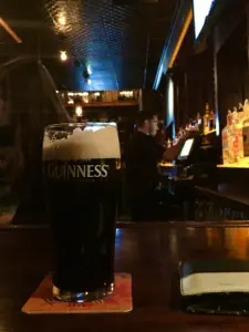 A pint of Guinness stout on a coaster sits on a bar counter with a wallet nearby; behind it, bartenders work and liquor bottles line the shelves in a dimly lit bar.