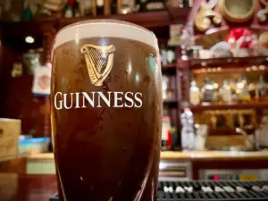 A close-up of a Guinness pint glass, showcasing the iconic logo and harp. The softly blurred background reveals a classic pub setting with shelves of bottles and ornate woodwork.