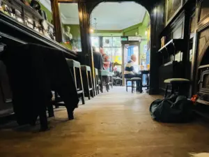 Low-angle view of a cozy pub interior with wooden floors, bar stools, and people sitting at tables near large windows. The atmosphere is warm, with green walls and dim lighting. A bag sits on the floor in the foreground.