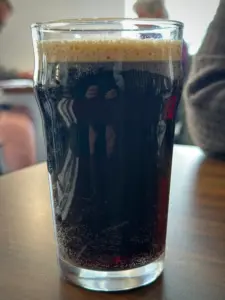 A pint glass filled with dark, frothy beer sits on a table. The glass has a layer of foam on top, and there are blurred figures and reflections in the background.