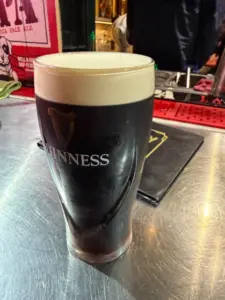 A pint glass of Guinness stout with a creamy head sits on a metal bar counter, with bar tools and drink mats visible in the background.