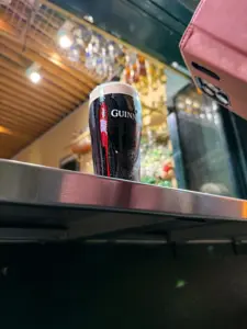 A pint of Guinness stout sits on a metal countertop in a bar, with glassware hanging above and a pink phone case partially visible in the upper right corner of the image.