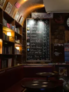 A cozy pub interior with a chalkboard menu listing draft beers, wooden shelves filled with books, red cushioned seating, and warm ambient lighting.