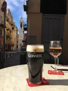 A Guinness pint glass and a glass of rosé wine sit on a round marble table outdoors at a pub, with colorful European buildings and a white clock tower in the background on a sunny day.