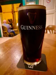 A Guinness pint glass sits on a coaster atop a wooden table in a pub, with people and warm lighting softly blurred in the background.