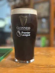 A Guinness pint glass with a creamy head sits on a wooden surface in a pub setting. The glass features the Guinness logo and the Premier League logo in white, with a softly blurred background.