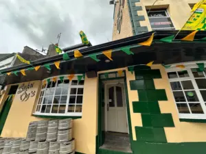 A pub with green and yellow bunting, green trim, and Guinness pint glasses visible inside. The building has rounded windows, a sign reading "Bar O’s," and kegs stacked outside under a cloudy sky.