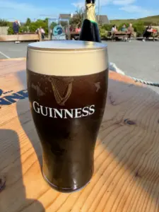 A Guinness pint glass sits on a wooden outdoor table in sunny weather, with a glass of Rockshore and people enjoying themselves at picnic benches in the background, creating a classic pub atmosphere.