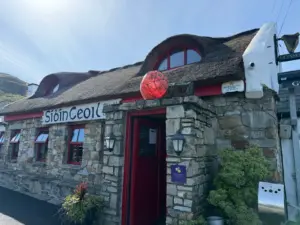 A rustic stone cottage with a thatched roof, red trim, and round windows. A large red buoy hangs above the entrance beside a Guinness pint glass sign. The sign reads "Si6inCeol," with shrubs visible outside.