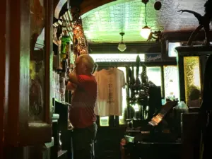 A bartender in a dimly lit pub fills a glass from a tap. A white T-shirt hangs behind the bar, and vintage decor and bottles line the shelves. Greenish light filters in from a window.