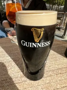 A pint of Guinness with a creamy head sits on an outdoor table in sunlight; a person holding a phone and a glass of orange-colored drink are visible in the background.