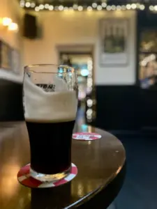A half-full Guinness pint glass with a foamy head sits on a coaster atop a polished wooden table in a softly lit pub, with blurred lights and wall decor visible in the background.