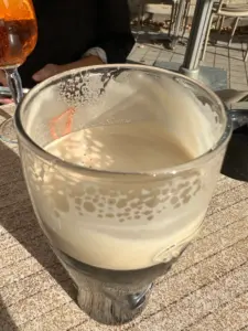 A partially finished pint of stout beer with a creamy head sits on a sunlit outdoor table. Another person is visible in the background holding a drink with an orange hue.