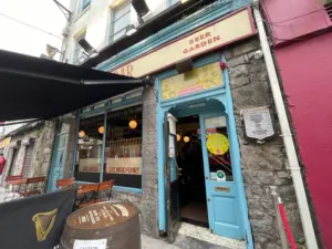 Street view of a pub with a stone facade, blue door, and "Beer Garden" signs. Outdoor seating features tables, chairs, and a Guinness pint glass atop a barrel. A partially open door offers a glimpse into the inviting interior.