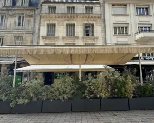 A street café with a large beige awning and planters filled with greenery in front, set against ornate stone buildings with large windows and balconies.