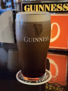 A Guinness pint glass with a creamy head sits on a coaster atop a dark pub bar counter, with a Guinness sign and orange background visible behind it.