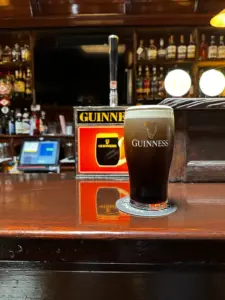 A Guinness pint glass sits on a coaster at a wooden pub bar, with a Guinness-branded tap and various bottles and bar equipment in the background.