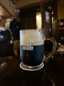 A Guinness pint glass with a foamy head sits on a coaster on a dark wooden bar, "CHANGING TIMES BREWERY" printed on the glass, in a cozy pub setting.
