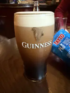 A freshly poured pint of Guinness with a creamy head sits on a wooden table next to a blue bag of Tayto crisps and an empty glass in a dimly lit pub.