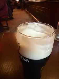 A pint glass of Guinness beer with a thick, creamy head sits on a wooden table in a dimly lit pub, with blurred patrons and bar in the background.