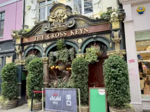 The ornate entrance of The Cross Keys pub features arched windows, gold detailing, potted plants, and greenery. Signs for Asahi beer and Budweiser stand outside, with trimmed bushes flanking the doorway.