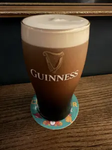 A Guinness pint glass with a creamy head sits on a colorful coaster atop a wooden table in a pub, with part of a dark wall and gold picture frame in the background.