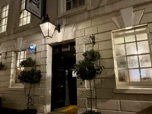 Nighttime photo of a white stone building with large windows, a black door under a lit street lantern, potted plants on either side of the entrance, and a hanging sign above the door that hints at a Guinness pint glass pub inside.