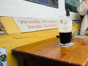 A Guinness pint glass rests on a small wooden table outside, beside a sign reading "World's Smallest Beer Garden" on a yellow wall. A person’s arm appears in the corner, adding to the cozy pub setting.