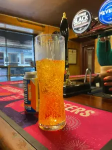 A tall glass of bright orange, fizzy drink with a straw stands on a bar counter next to a can of Irn-Bru Sugar Free. Beer taps and a person’s hand are visible in the background.