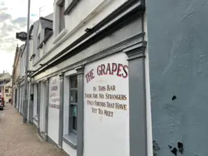 A street view of a grey and white building with a sign reading, “THE GRAPES. In this bar there are no strangers, only friends that have yet to meet.” The sidewalk runs alongside the building under a cloudy sky.