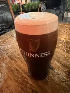 A Guinness pint glass with a creamy head sits on a wet wooden table at an outdoor pub setting, with blurred greenery and a chalkboard in the background.