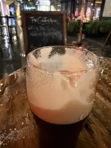 A partially finished pint of Guinness sits on a wet wooden table outdoors at night, with a chalkboard menu and blurred lights in the background at the pub. Rain glistens on the table and ground.